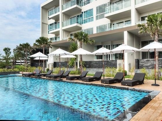 Poolside at a modern hotel with black lounge chairs, white umbrellas, and palm trees along the edge of a blue tile swimming pool.