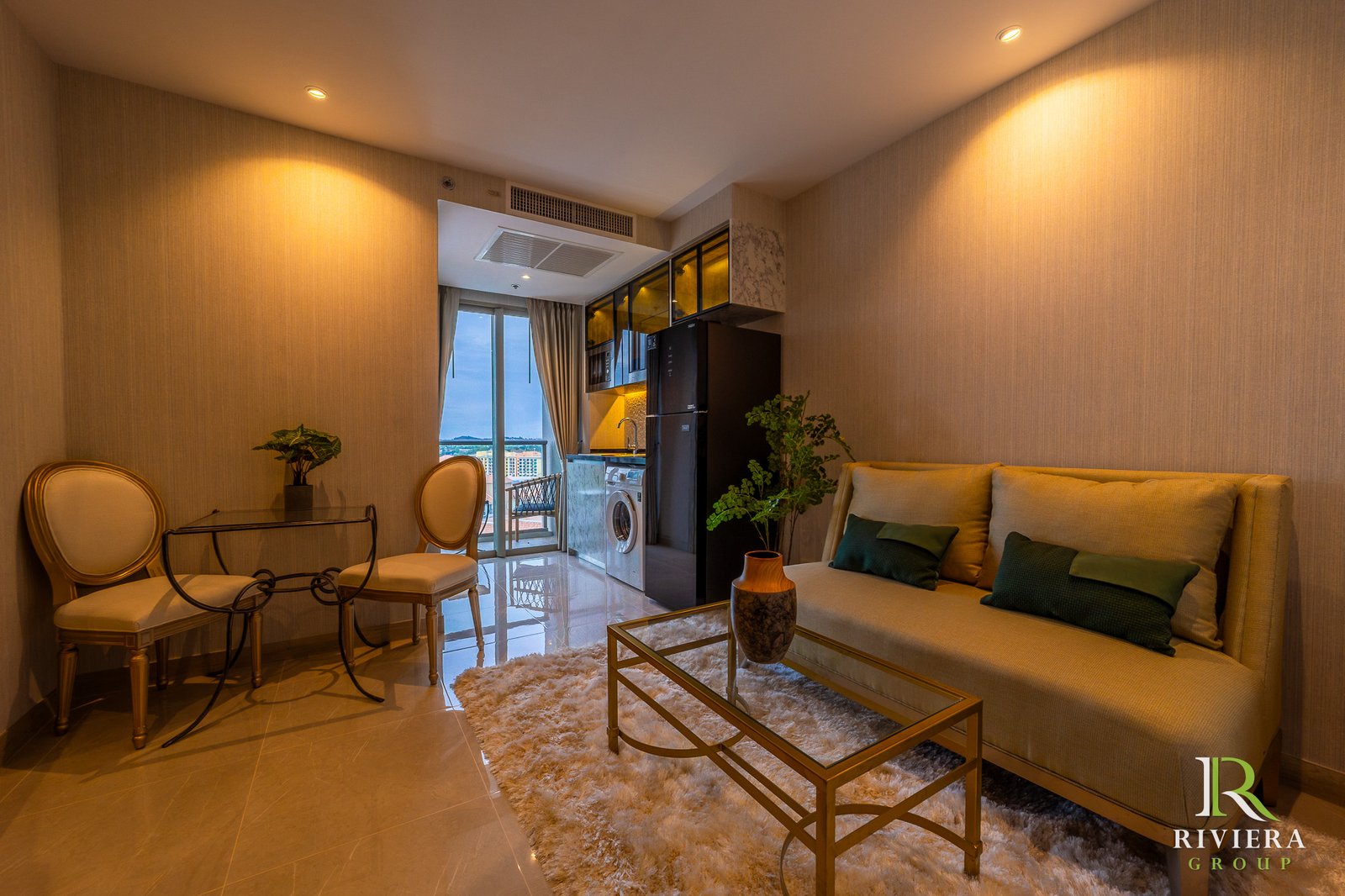 Open-plan living area with beige sofa, glass coffee table, two dining chairs, and a kitchen visible through an open doorway; warm lighting and a fluffy rug dominate the scene.