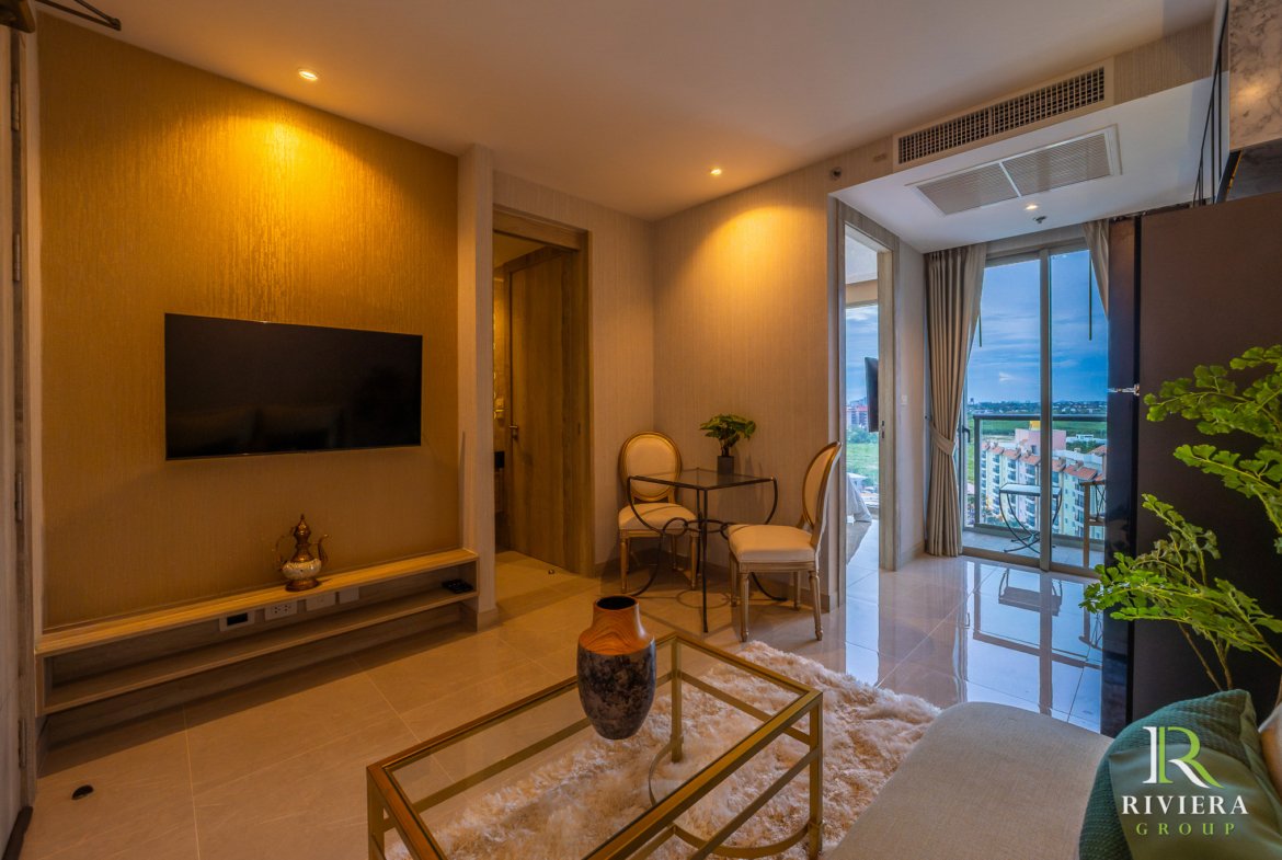 Modern living room with a wall-mounted TV, glass coffee table, beige seating, and a balcony view of buildings outside.