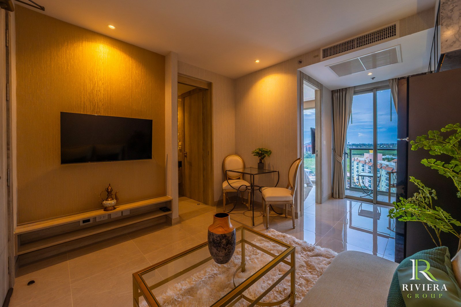 Modern living room with a wall-mounted TV, glass coffee table, beige seating, and a balcony view of buildings outside.