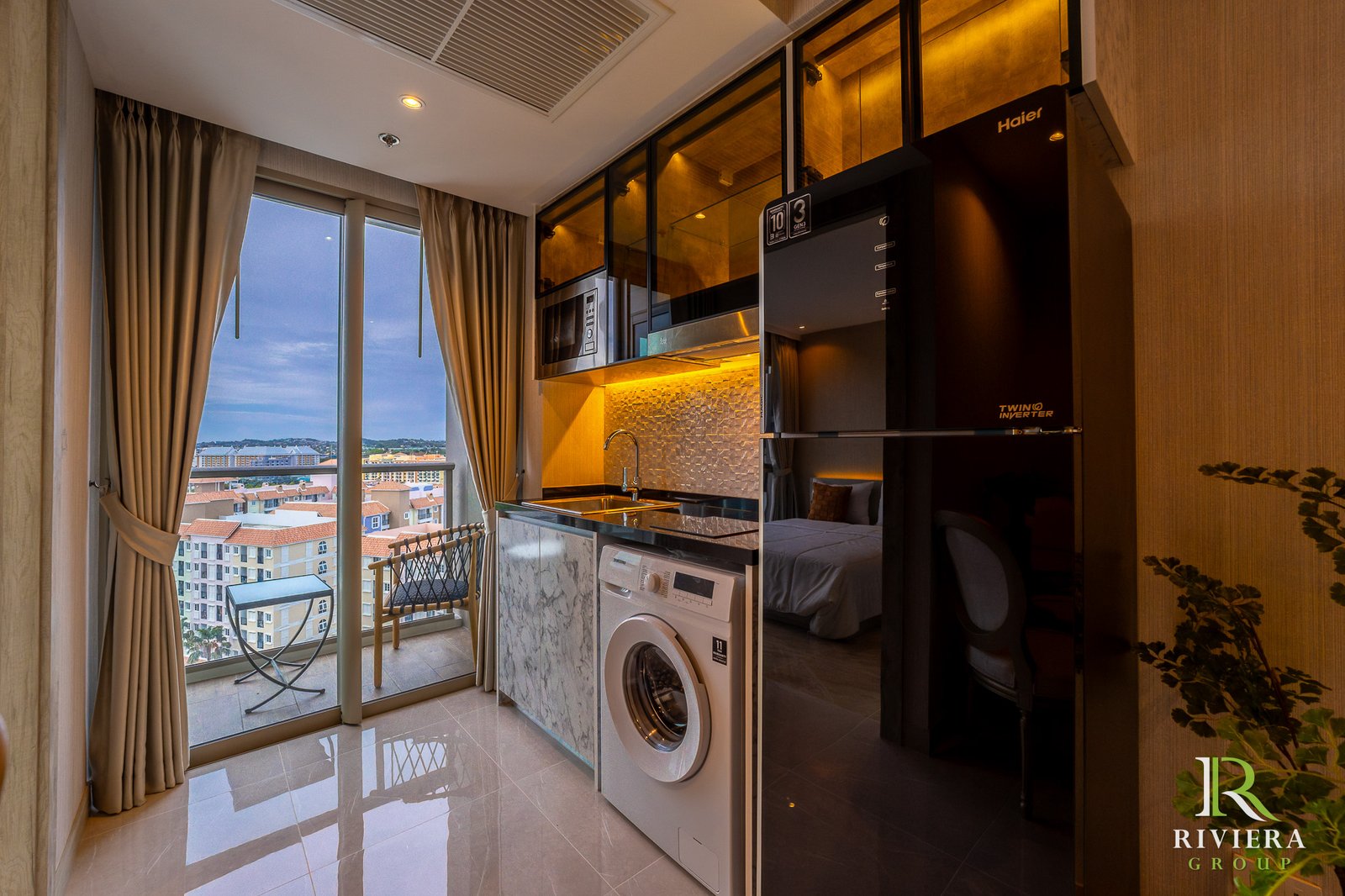 Modern kitchen with built-in microwave, fridge and washing machine beside glass balcony doors overlooking a cityscape.