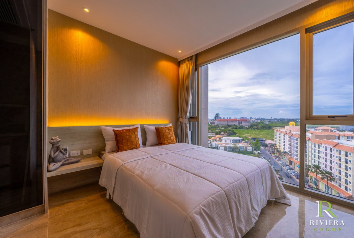 Hotel room with a double bed, white linens, orange throw pillows, and warm wall lighting beside a large window overlooking the cityscape.