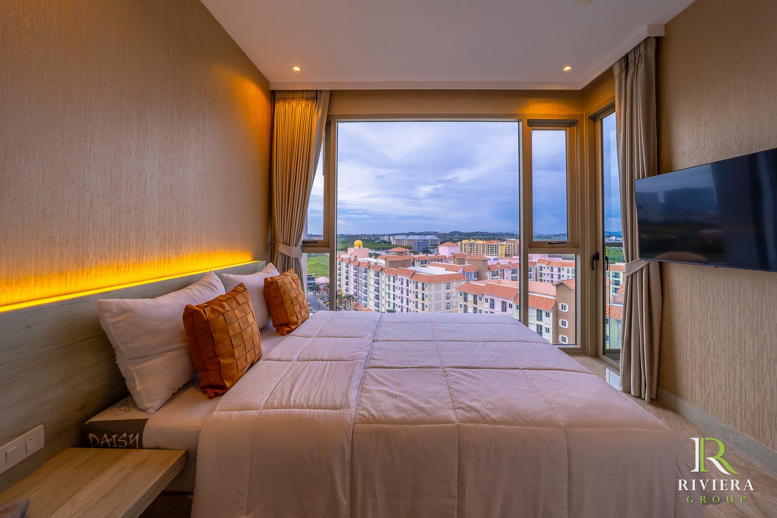 Hotel bedroom with warm backlit headboard, beige bedding, and orange plaid pillows, large window overlooking a cityscape and blue sky, wall-mounted TV to the right.