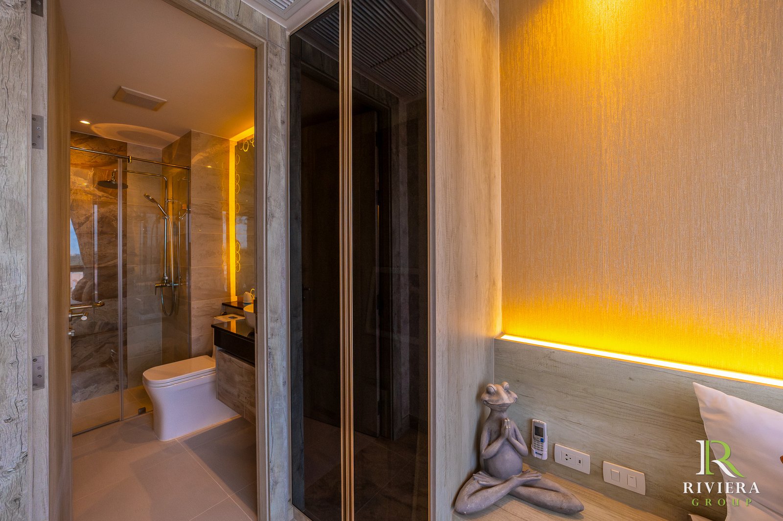 Luxurious hotel bathroom with glass shower, marble walls, and warm amber lighting; decorative sculpture on the shelf and Riviera Group logo in corner.