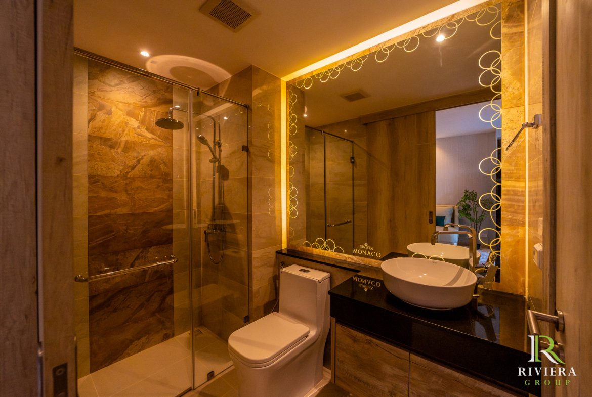 Luxurious hotel bathroom with glass shower enclosure, white toilet, dark vanity, and a circular-pattern mirror reflecting warm lighting; Riviera Monaco branding visible on the counter surface.