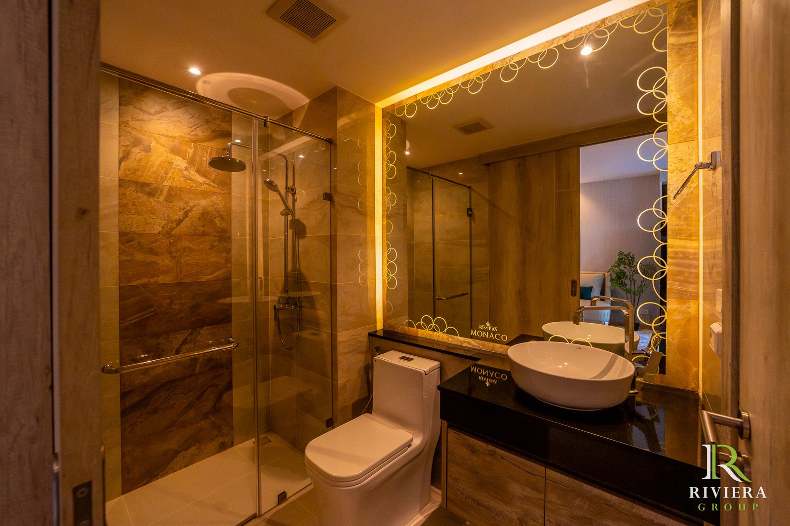 Luxurious hotel bathroom with glass shower enclosure, white toilet, dark vanity, and a circular-pattern mirror reflecting warm lighting; Riviera Monaco branding visible on the counter surface.