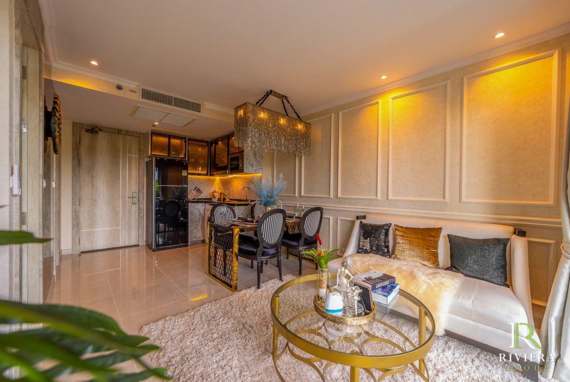 Open-plan living area with cream walls, a gold crystal chandelier, beige sofa with pillows, and a glass coffee table; kitchen area in the background.