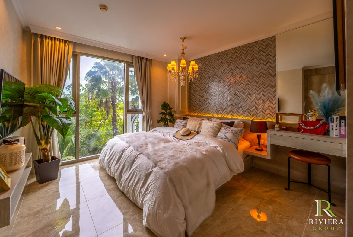 Luxurious bedroom with a king bed, beige bedding, and a crystal chandelier, overlooking tropical plants through large glass doors.