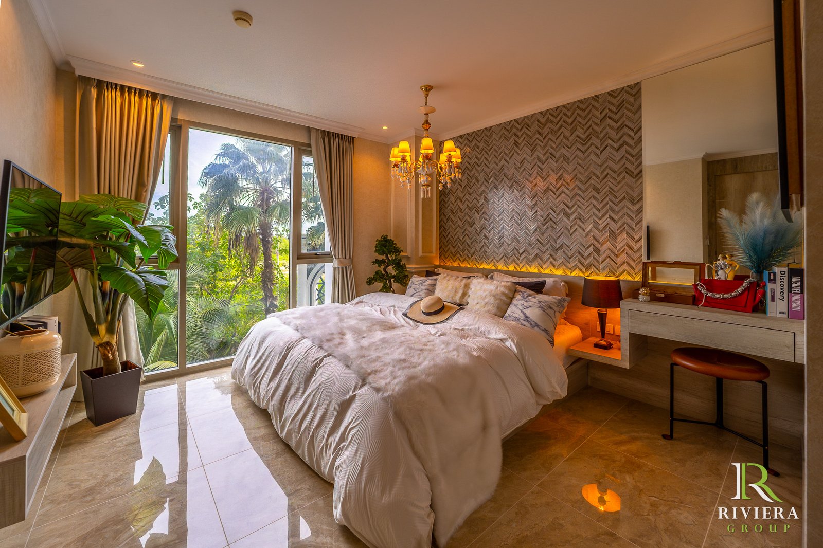 302-S10 Luxurious bedroom interior with a chandelier, king bed, plush pillows, large windows and plants in a sunlit suite. Branding visible: Riviera Group.