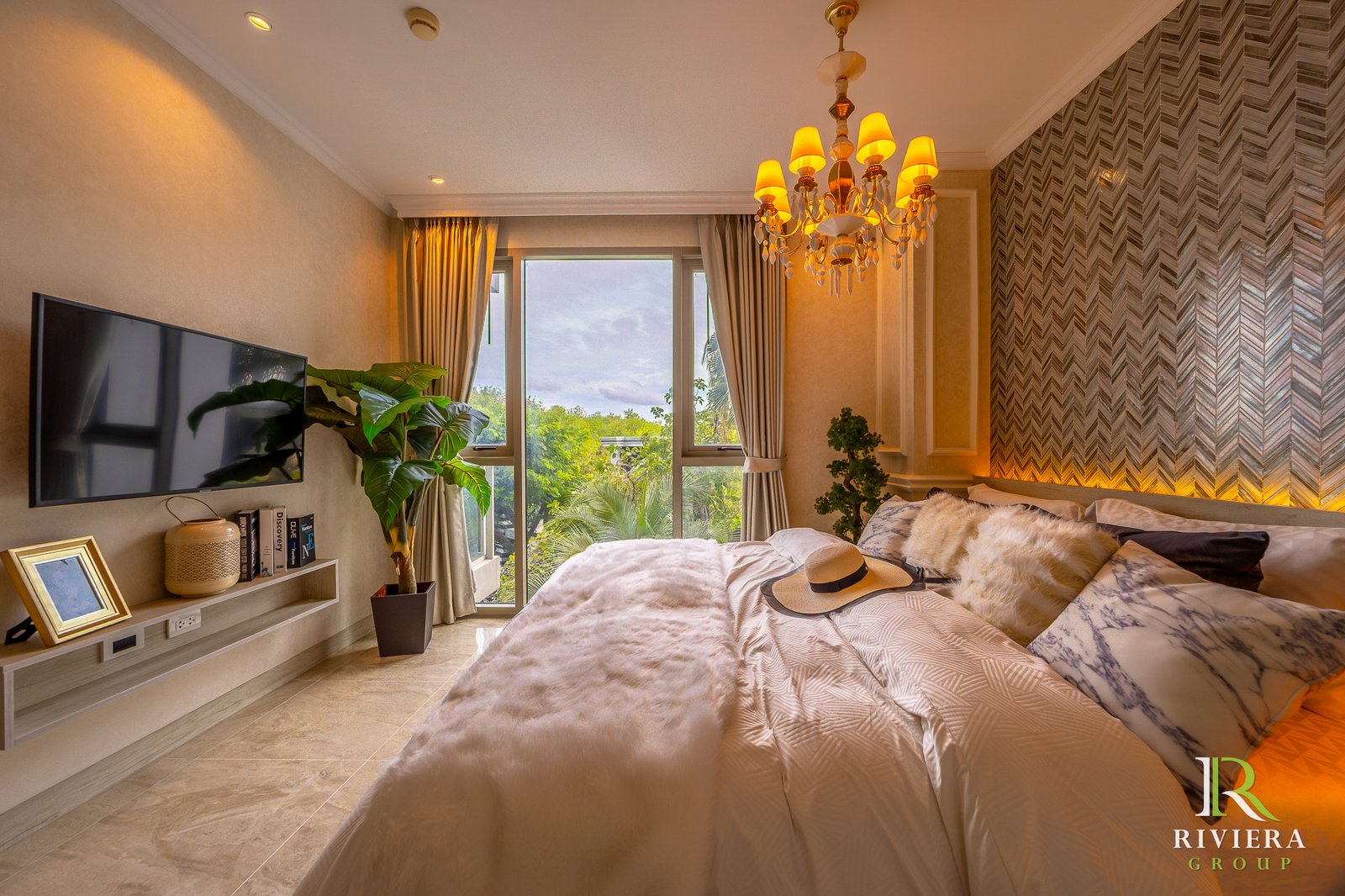 302-S11 Luxurious bedroom with beige walls, a crystal chandelier, large window with curtains, plush bedding, and a wall-mounted TV on a shelf beside tropical plants in front of a chevron accent wall.