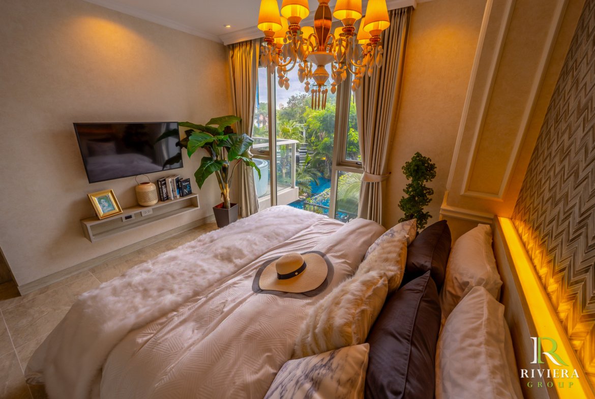 Luxurious hotel bedroom with a chandelier, beige walls, a plush bed, and a large window overlooking greenery outdoors.