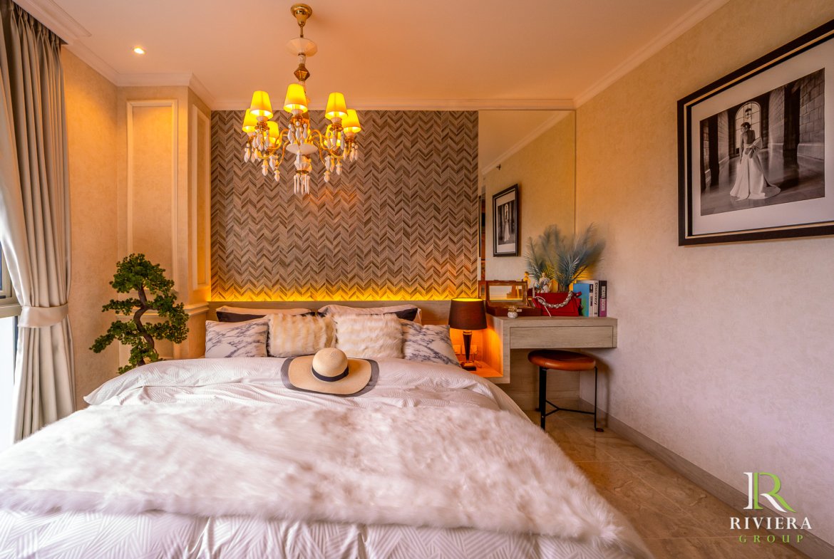 Hotel-style bedroom with a crystal chandelier, chevron accent wall, and two beds topped with fur throws and a straw hat. A large framed black-and-white photo hangs on the right wall; a desk with books and a small plant sits by the window. Riviera Group logo visible bottom-right.
