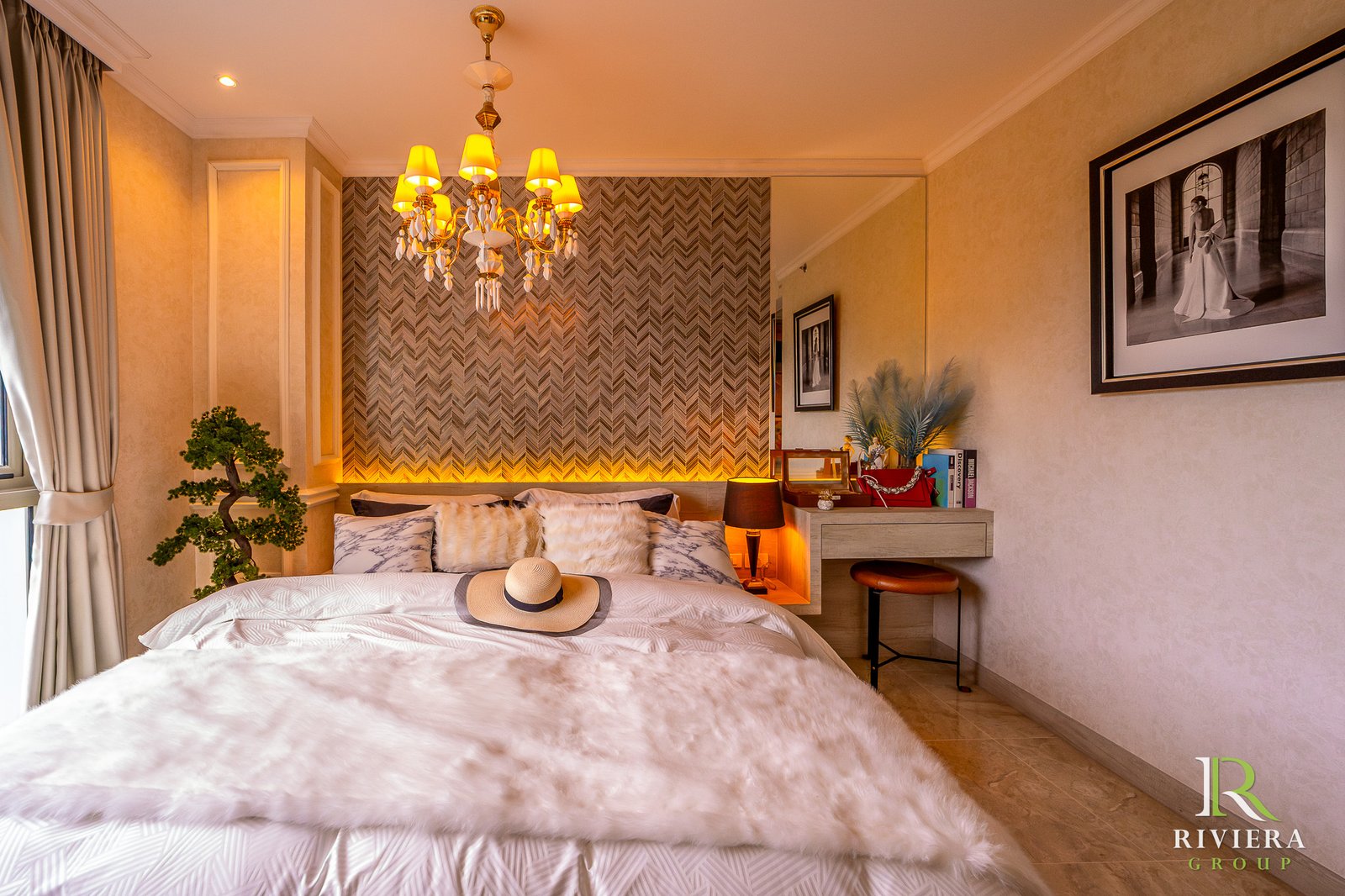 302-S14 Hotel-style bedroom with a crystal chandelier, chevron accent wall, and two beds topped with fur throws and a straw hat. A large framed black-and-white photo hangs on the right wall; a desk with books and a small plant sits by the window. Riviera Group logo visible bottom-right.
