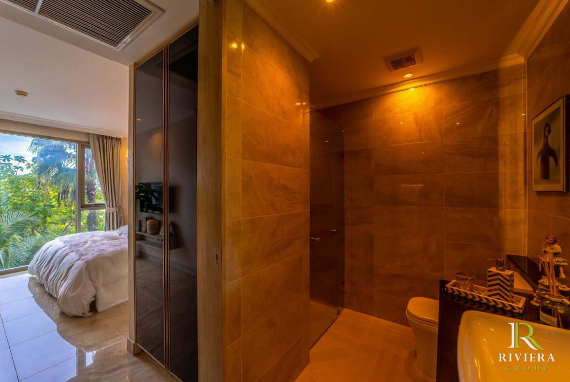Marble-tiled bathroom with a glass-walled shower, opening into a sunlit bedroom with a window view of trees and plants.