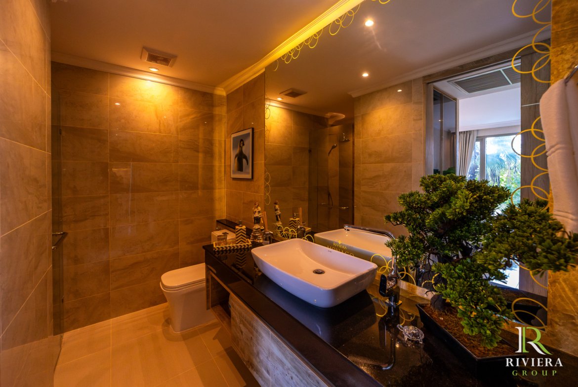 Luxurious bathroom with double white vessel sinks on a dark counter, beige tiled walls, and a large mirror reflecting warm lighting. Green plants sit by the sink. RivierA Group logo in the lower right corner.