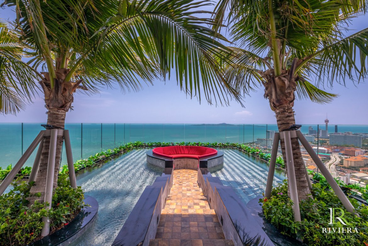Rooftop pool with a red circular seating area, palm trees, and a glass barrier overlooking a coastal city skyline.