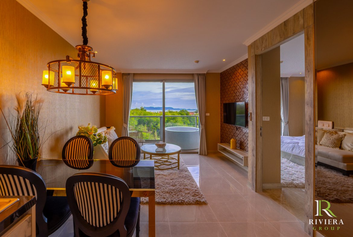 Hotel suite living and dining area with a round metal chandelier, glass dining table, striped chairs, and a balcony view of the ocean and greenery outside.