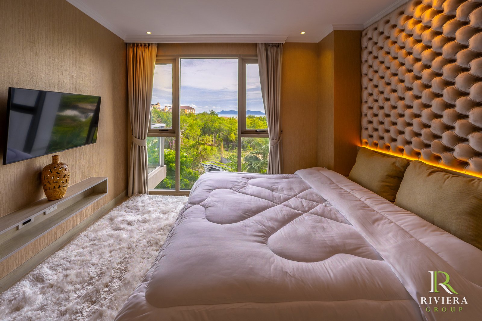 502-S10 Luxurious hotel bedroom with a tufted beige headboard, wall-mounted TV, and a large window offering greenery and distant buildings outside.