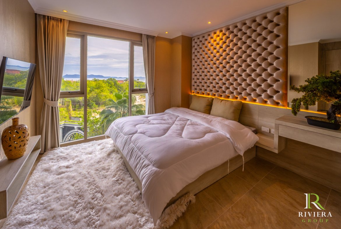 Luxurious hotel bedroom with a low platform bed, white duvet, and a large tufted beige headboard; windows show greenery and distant water.