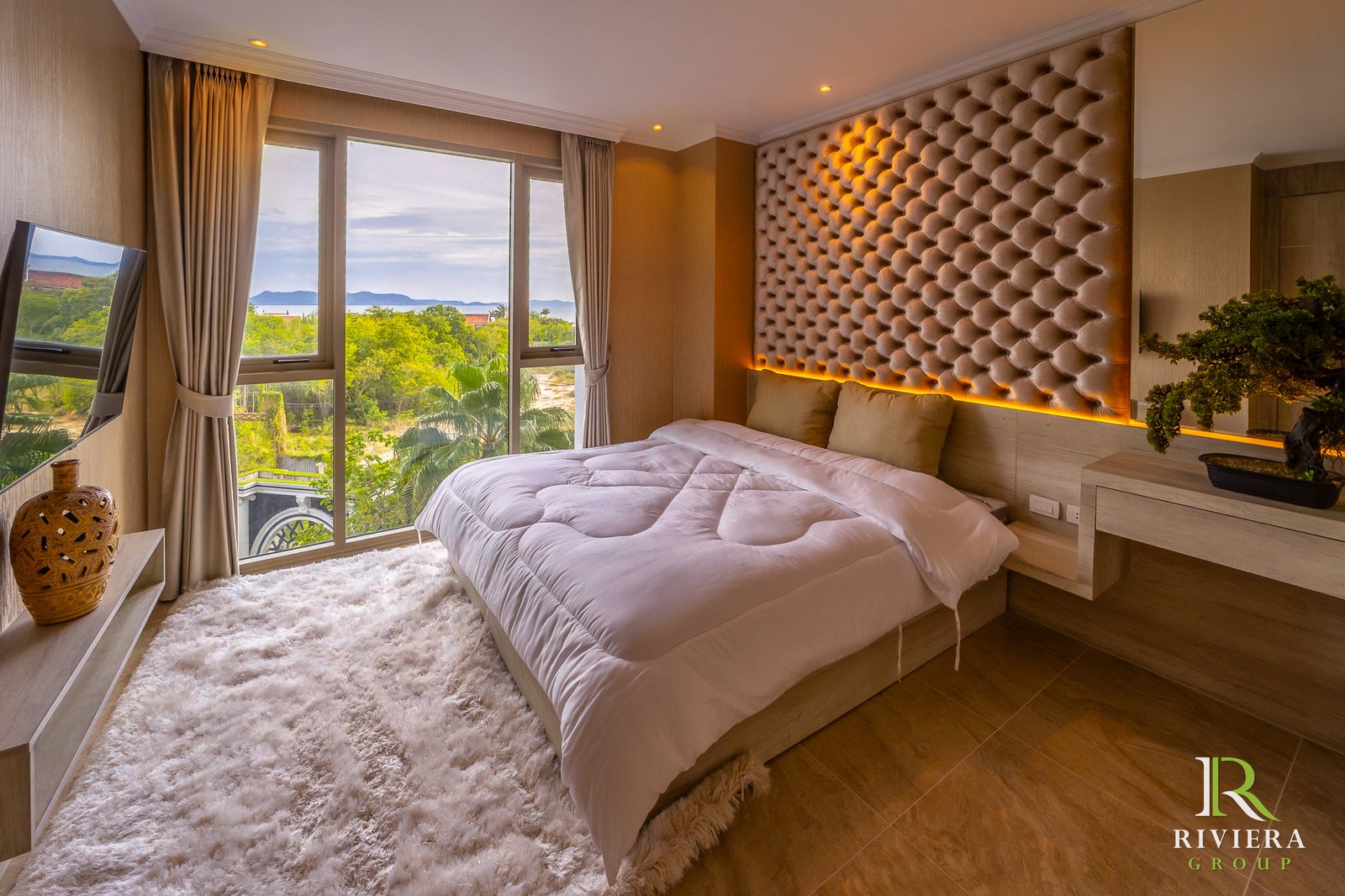 502-S11 Luxurious hotel bedroom with a low platform bed, white duvet, and a large tufted beige headboard; windows show greenery and distant water.