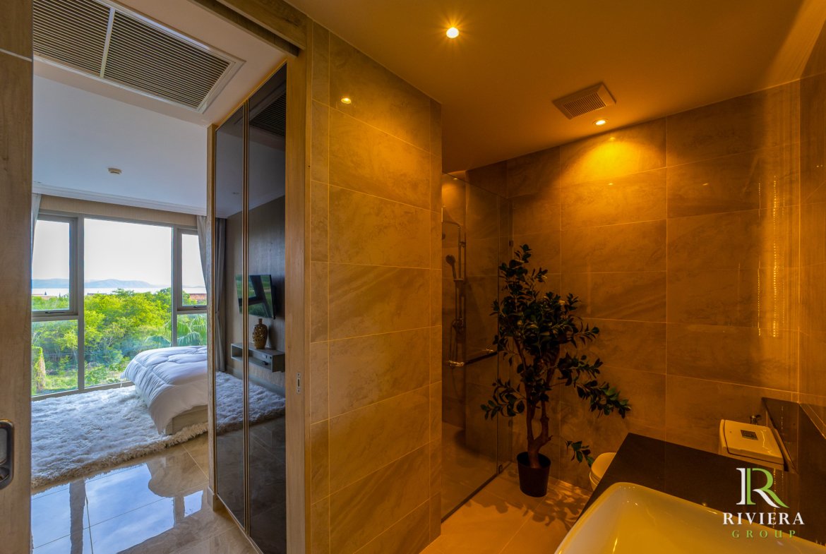 Modern hotel bathroom with marble walls and a glass-enclosed shower; doorway opens to a bright bedroom with a large window and greenery outside.