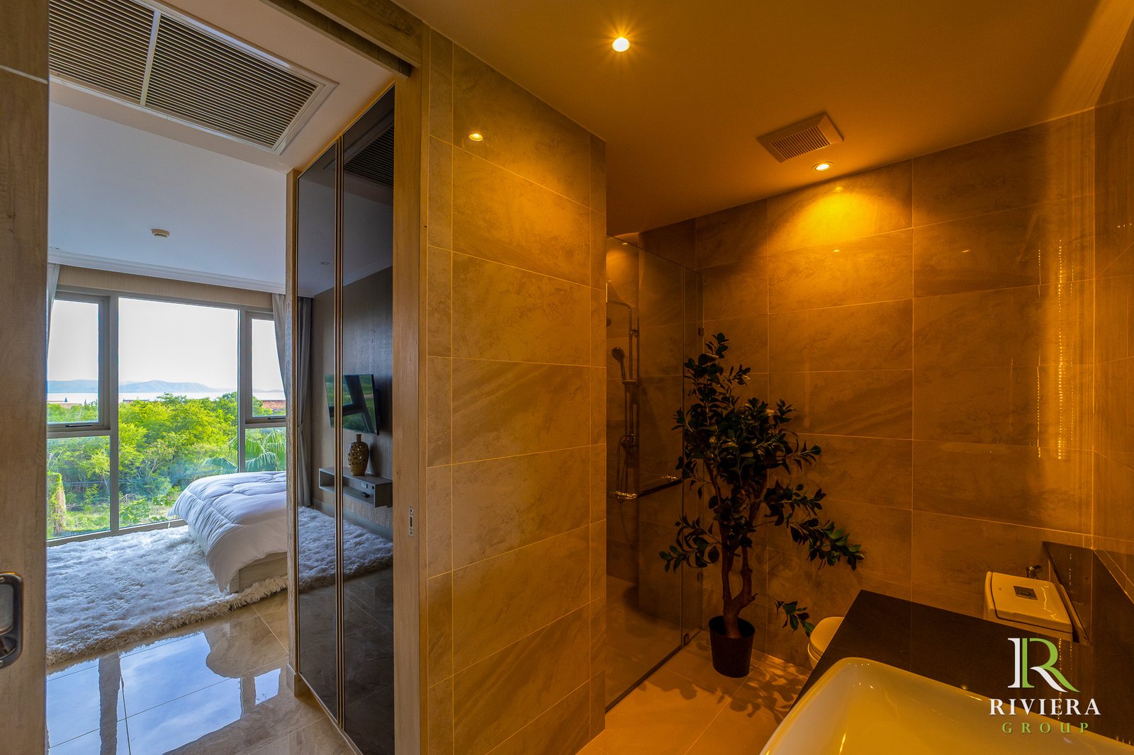 502-S15 Modern hotel bathroom with marble walls and a glass-enclosed shower; doorway opens to a bright bedroom with a large window and greenery outside.