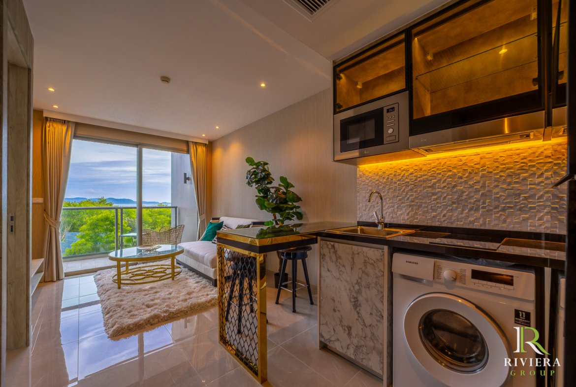 Open-plan living area with a modern kitchen featuring marble countertops and gold accents, a cream sofa, and a balcony with a hillside view.