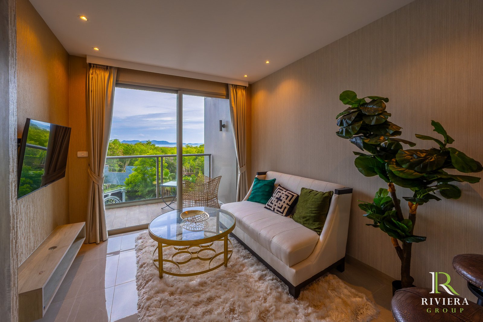 505-S03 Hotel-style living room with beige sofa, round glass coffee table, and a large potted plant; sliding doors open to a balcony with a scenic sea view.