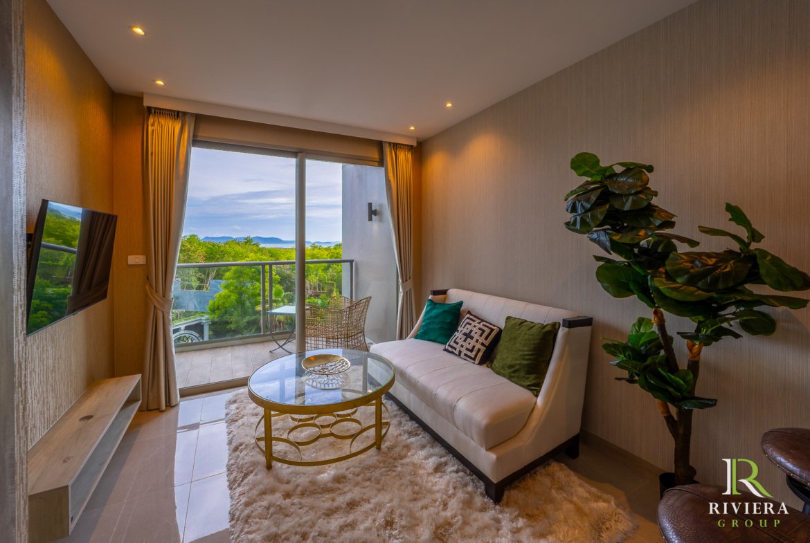 Living room with a beige sofa, glass coffee table on a fluffy rug, and large sliding doors opening to a balcony with trees and distant hills outside.