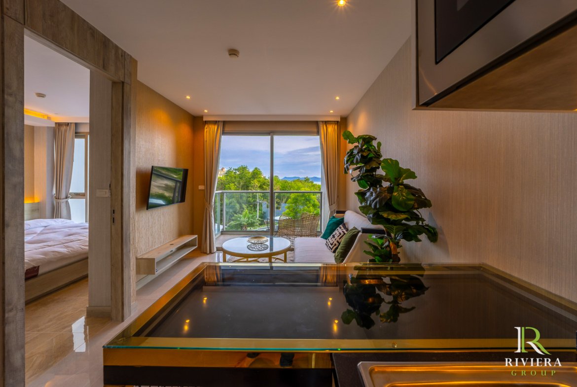 Spacious hotel living area with a glass coffee table, sofa, large windows, and a balcony view of trees and hills; indoor plant in foreground; Riviera Group logo bottom right