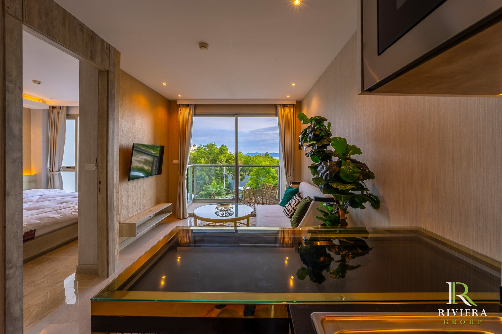 505-S04 Spacious hotel living area with a glass coffee table, sofa, large windows, and a balcony view of trees and hills; indoor plant in foreground; Riviera Group logo bottom right