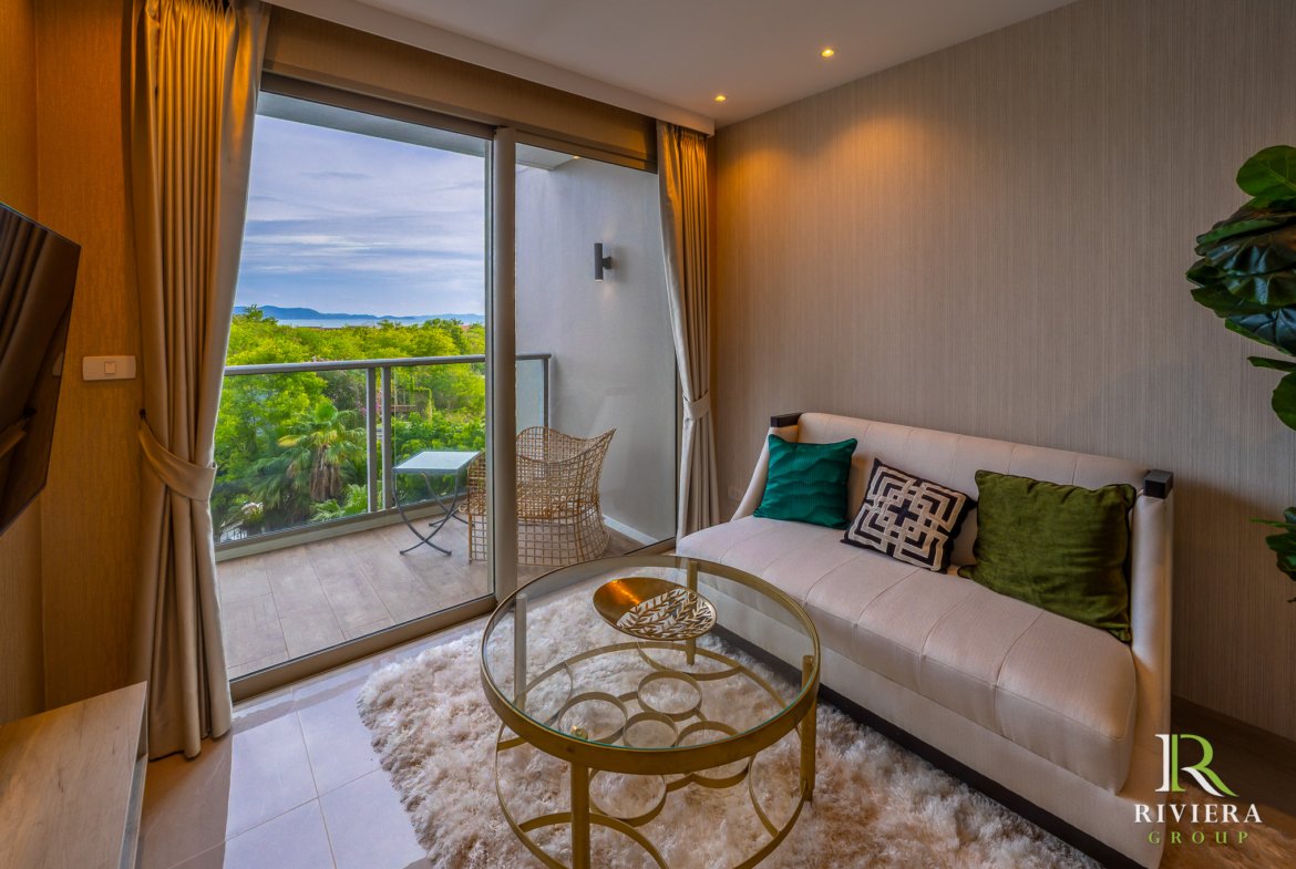 Living room with a beige sofa and colorful cushions, a round glass coffee table on a white shag rug, and a balcony view of trees and distant water.