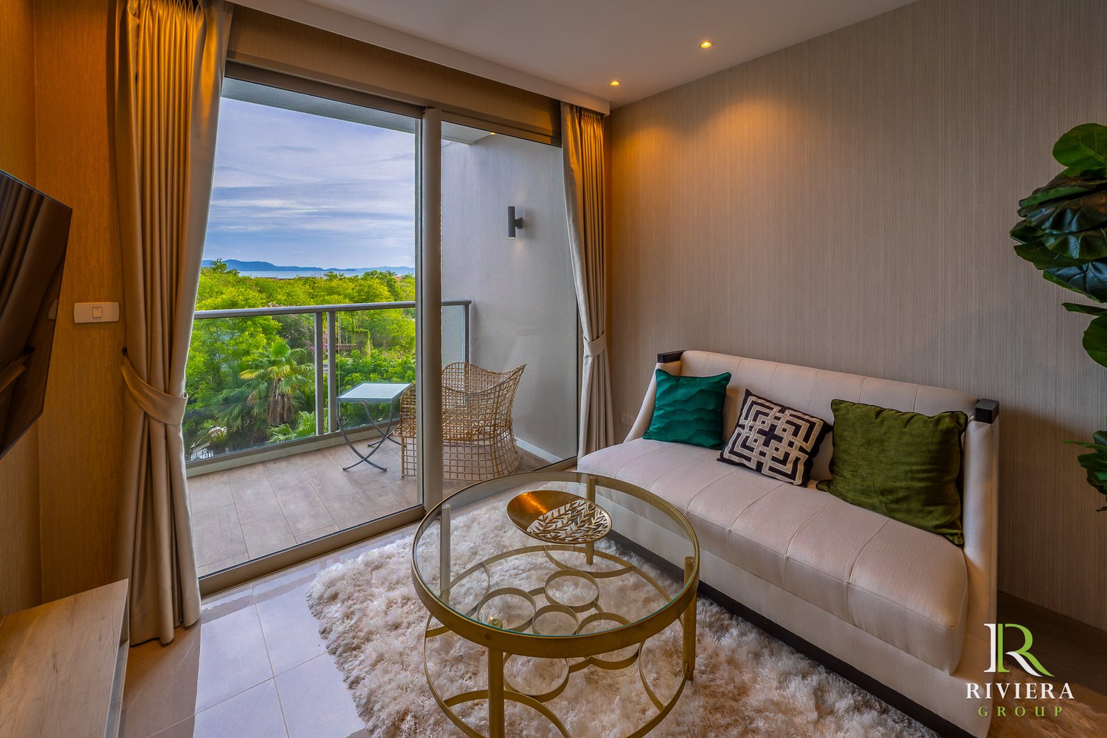 Living room with a beige sofa and colorful cushions, a round glass coffee table on a white shag rug, and a balcony view of trees and distant water.