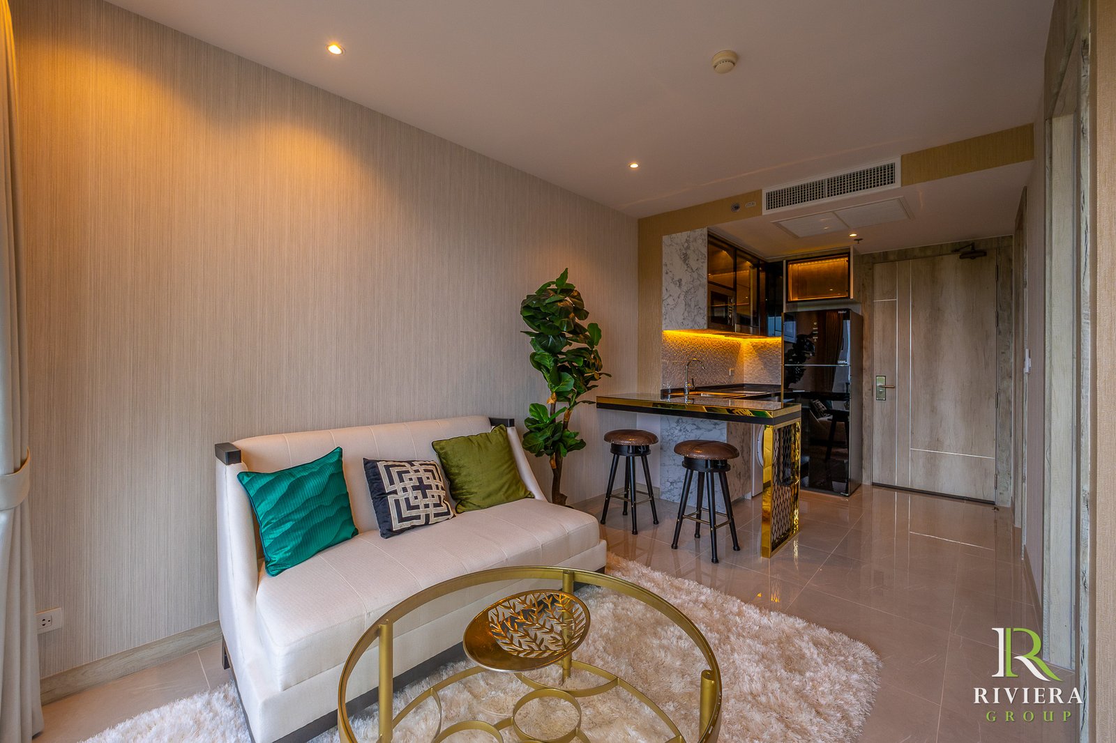 Contemporary living area with white sofa and teal/patterned cushions, a gold-framed glass coffee table, and a plant beside an open kitchen. A warm, upscale vibe.