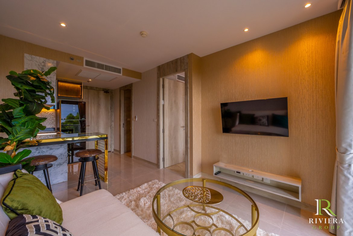 Modern living room interior with a wall-mounted TV, tan textured walls, and a glass coffee table on a fluffy white rug; warm recessed ceiling lights and a green plant add color to the space.