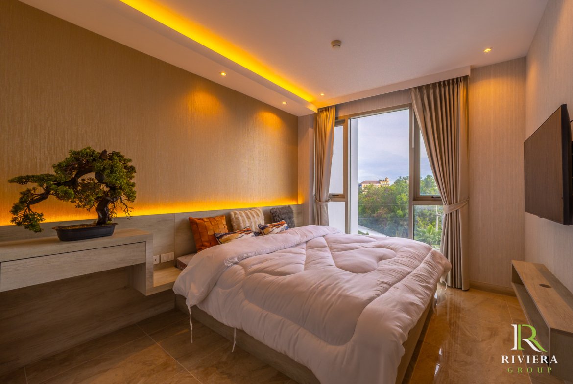 Hotel bedroom with two fluffy white beds, warm wood tones, and a bonsai on a dresser under yellow ambient lighting; window with beige curtains and a city view; Riviera Group logo in corner.
