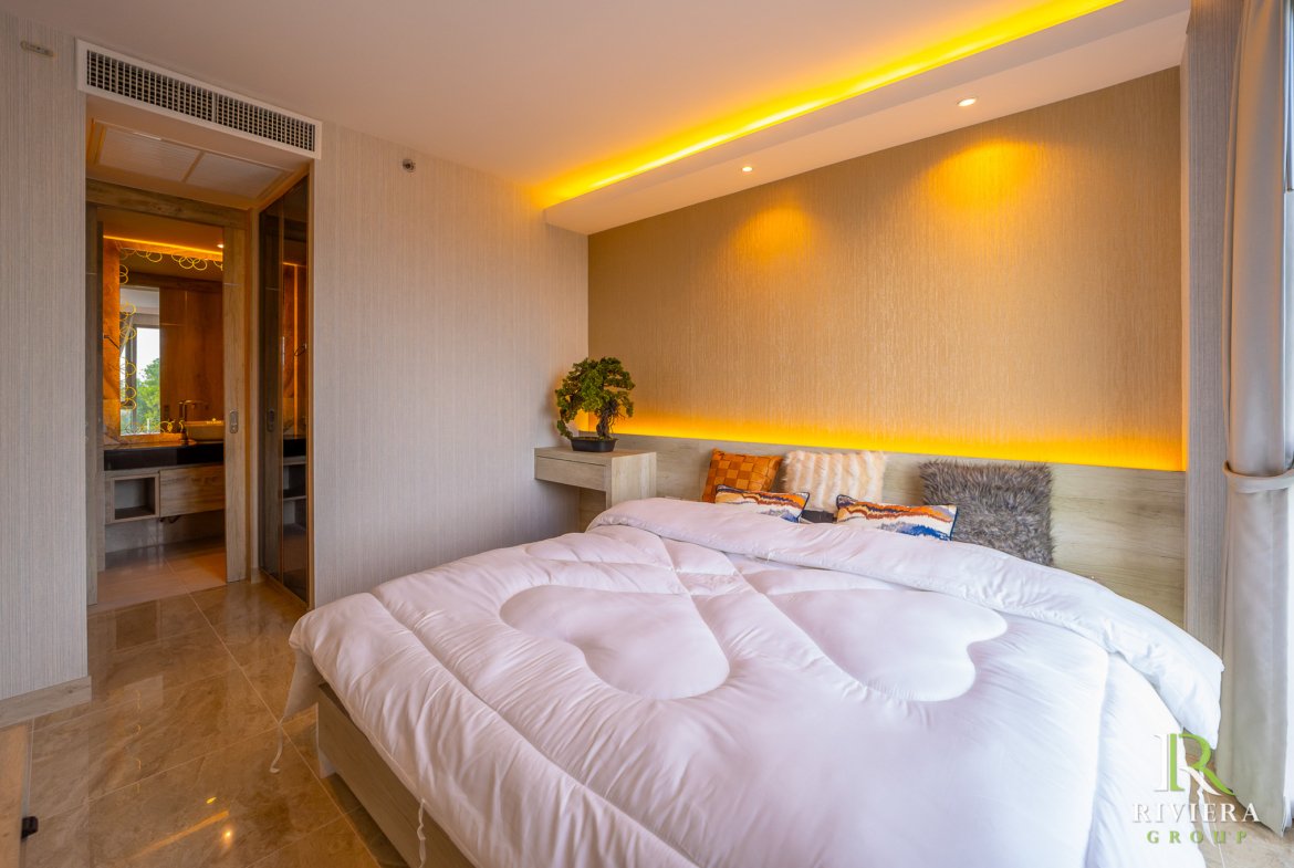 Hotel bedroom with two white-tufted beds, decorative cushions, and warm amber cove lighting along the headboard wall; a small plant on a nightstand and a doorway to the bathroom visible on the left.