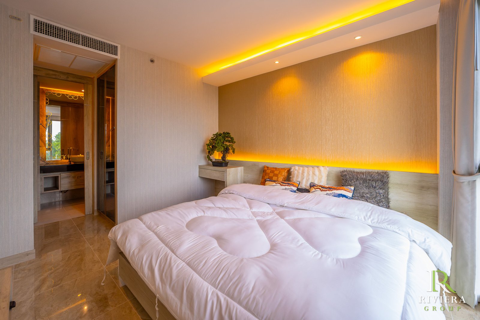Hotel bedroom with two white-tufted beds, decorative cushions, and warm amber cove lighting along the headboard wall; a small plant on a nightstand and a doorway to the bathroom visible on the left.