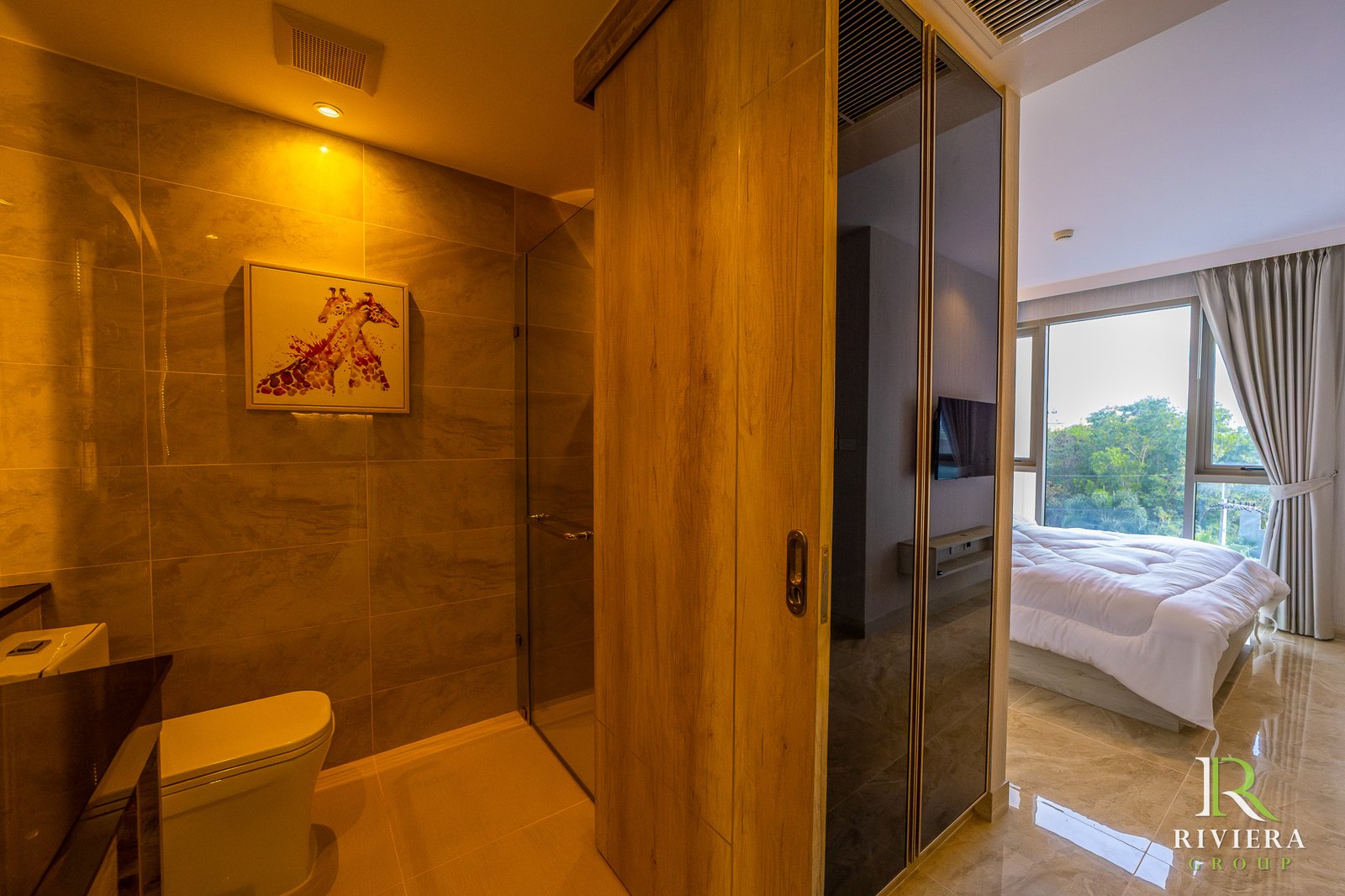 Hotel suite view: bathroom with beige tiles, framed giraffe artwork, and a wooden divider leading to a bedroom with a large window and white bed.