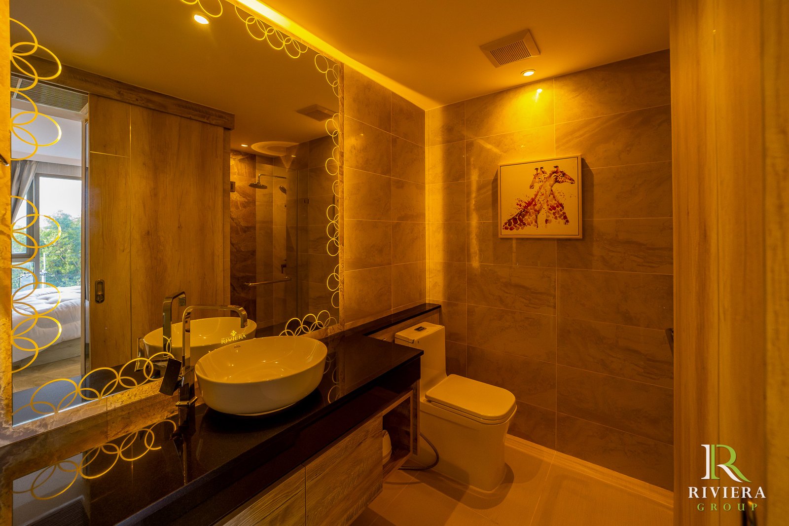 505-S15 Luxurious beige marble bathroom with a circular-pattern mirror, vessel sink, glass shower, and Riviera Group logo in the bottom right corner—warm yellow lighting.