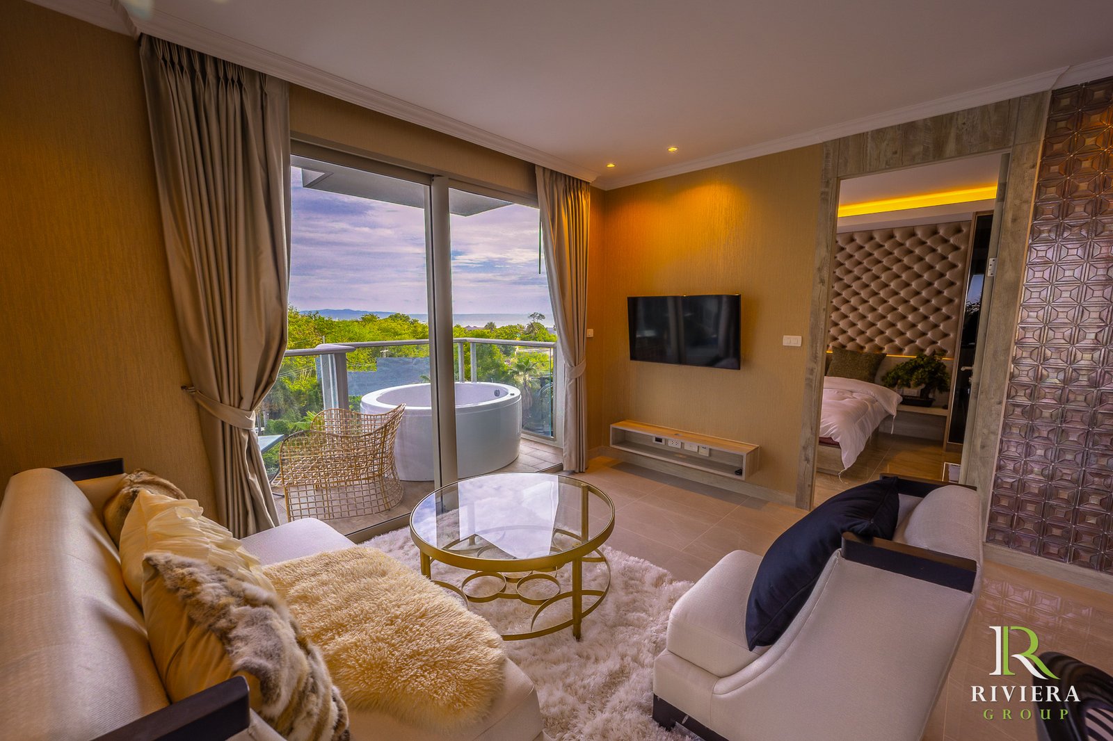 Modern living room with cream sofa, glass coffee table, and fluffy rug; large glass doors open to a balcony with a sea view. A wall-mounted TV and warm lighting enhance the cozy space.