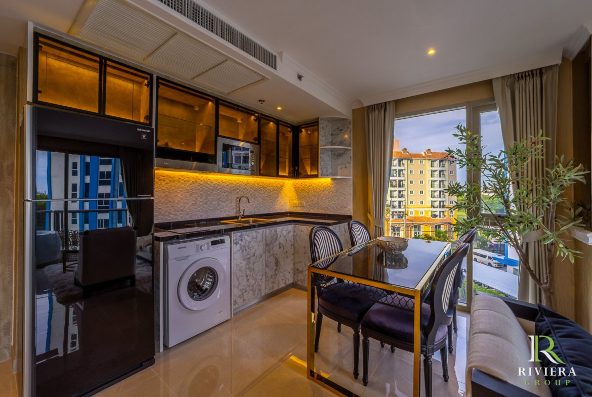 Modern kitchen with marble counters and warm under-cabinet lighting, dining table, and a large window showing an apartment building outside.