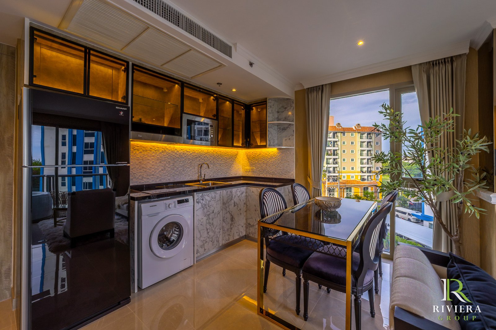 Modern kitchen with marble countertops, black appliances, and a glass dining table near a large window overlooking buildings outside.