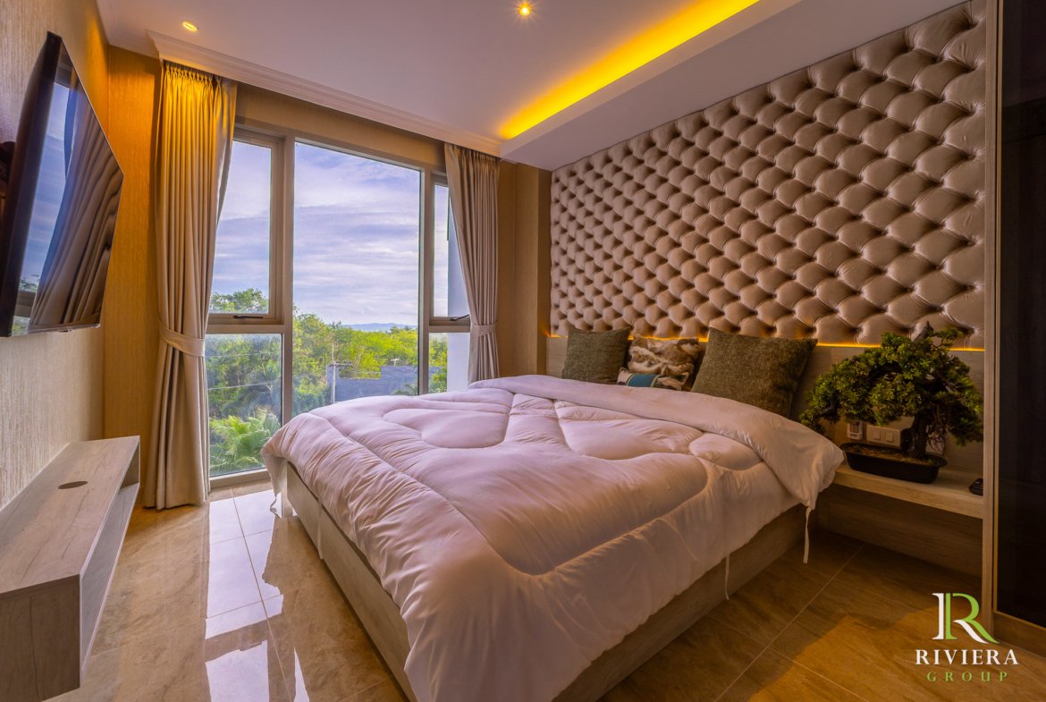 Luxurious hotel bedroom with a tall tufted beige headboard, white bedding, and large windows overlooking trees outside.