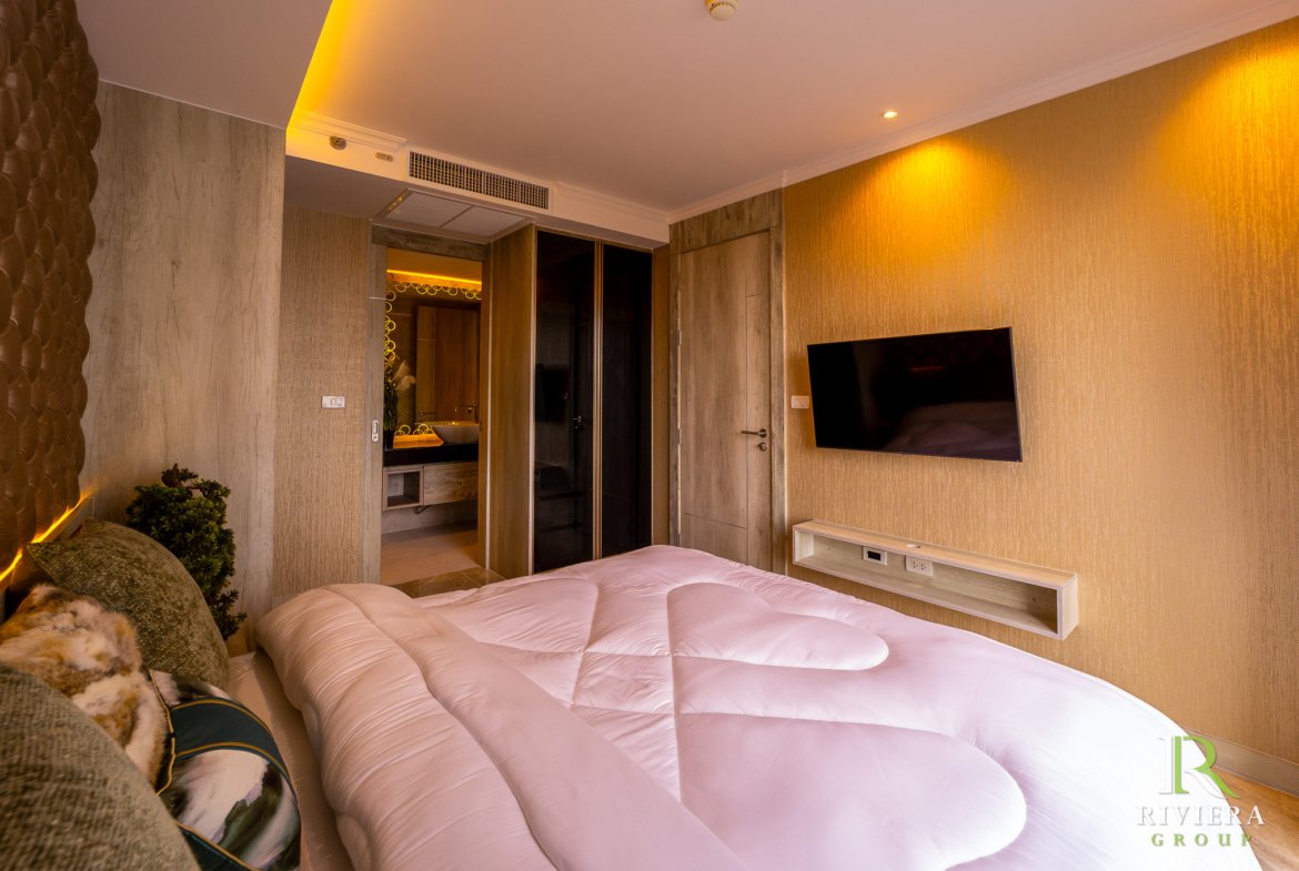 Hotel room with two white beds in the foreground, warm lighting, wall TV on a textured wall, and an open doorway revealing a sink area in the bathroom.