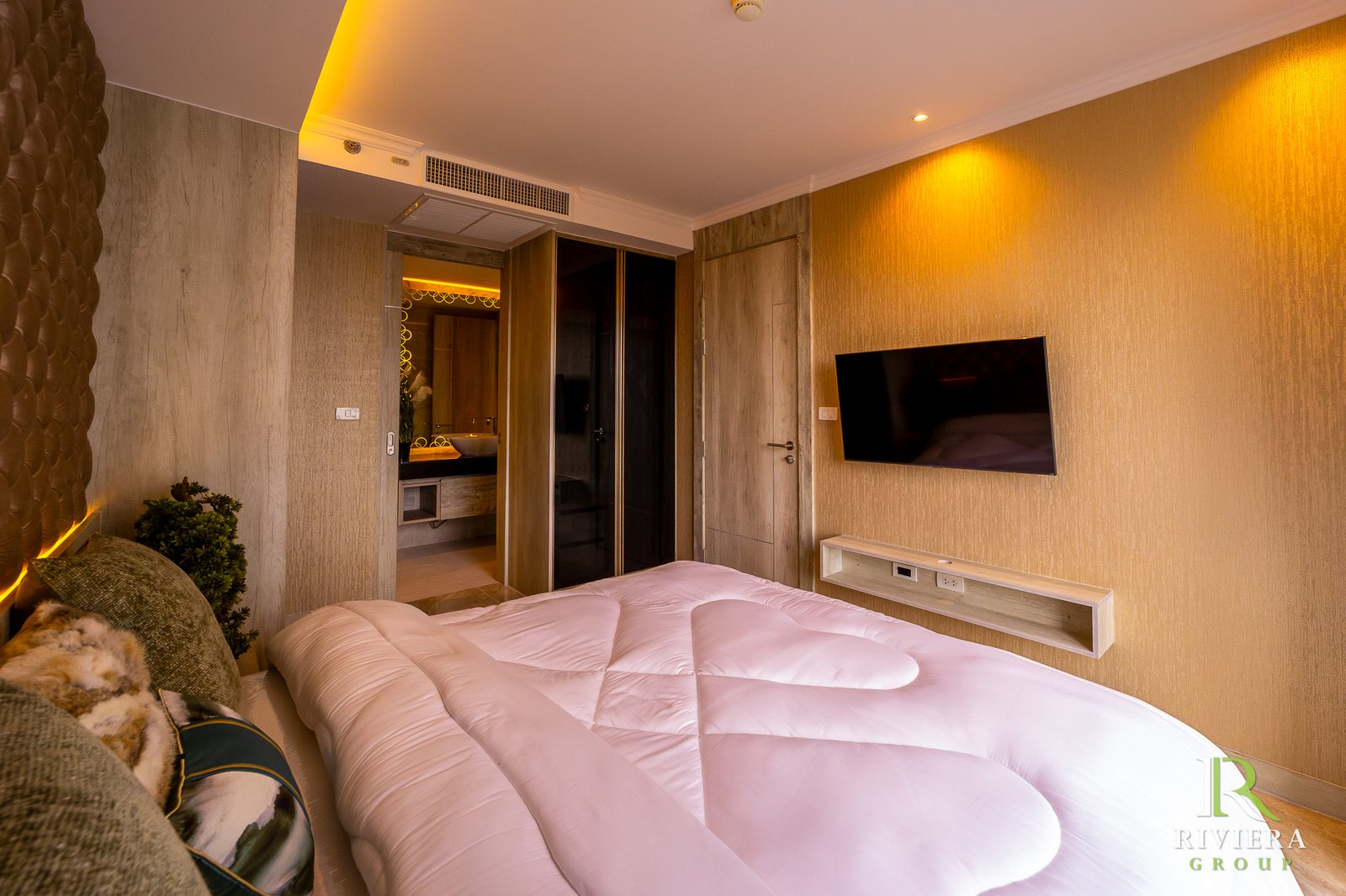 Hotel room with two white beds in the foreground, warm lighting, wall TV on a textured wall, and an open doorway revealing a sink area in the bathroom.