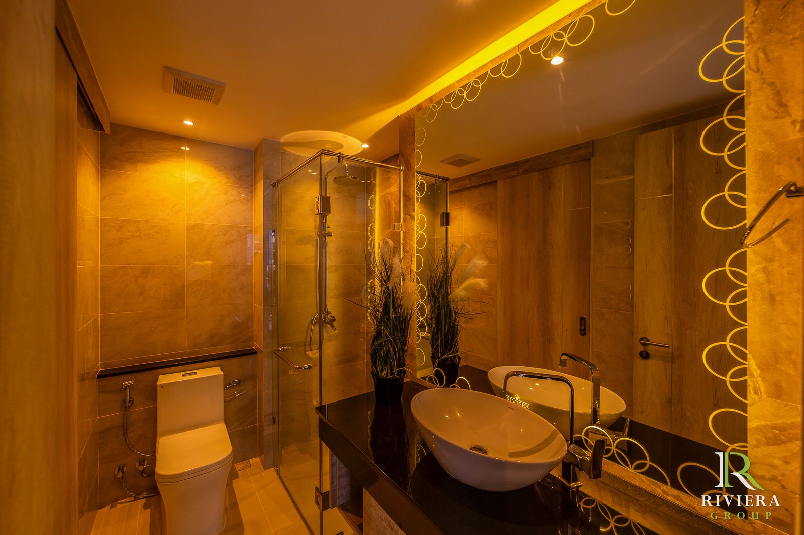 Luxurious bathroom with beige marble tiles, glass shower, twin vessel sinks, and Riviera Group logo in the lower right corner.