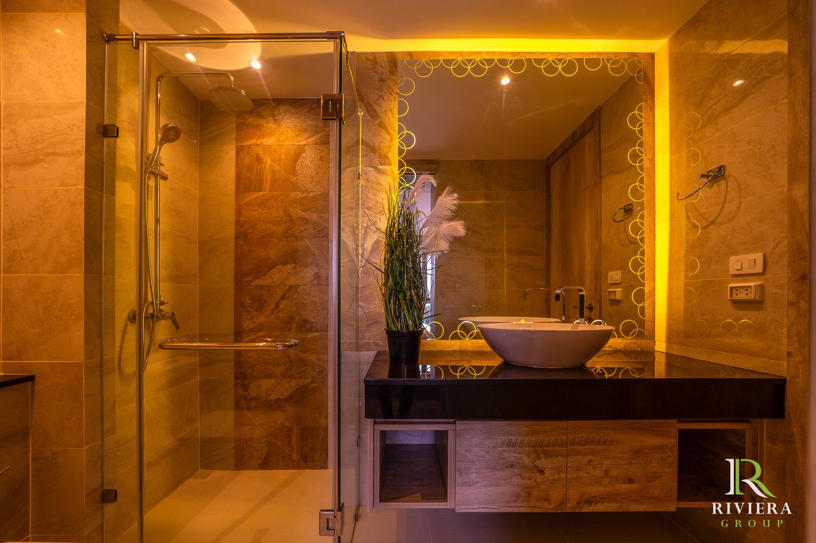 Modern marble bathroom with glass shower, vessel sink, tall plant, and a circular-ring mirror; warm amber lighting.