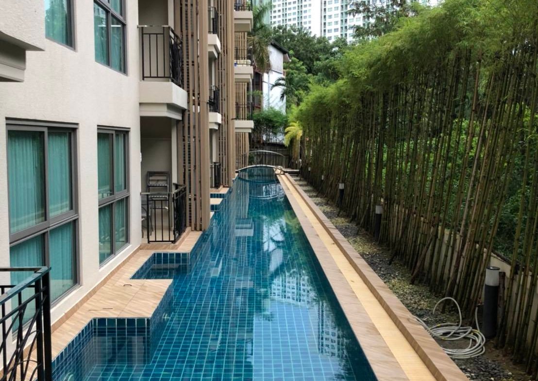 Narrow rectangular swimming pool with blue-tiled deck between a beige apartment building and a bamboo fence, with high-rise buildings in the distance.
