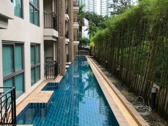Narrow rectangular swimming pool with blue tiled deck between a beige apartment building and a bamboo fence with high rise buildings in the distance